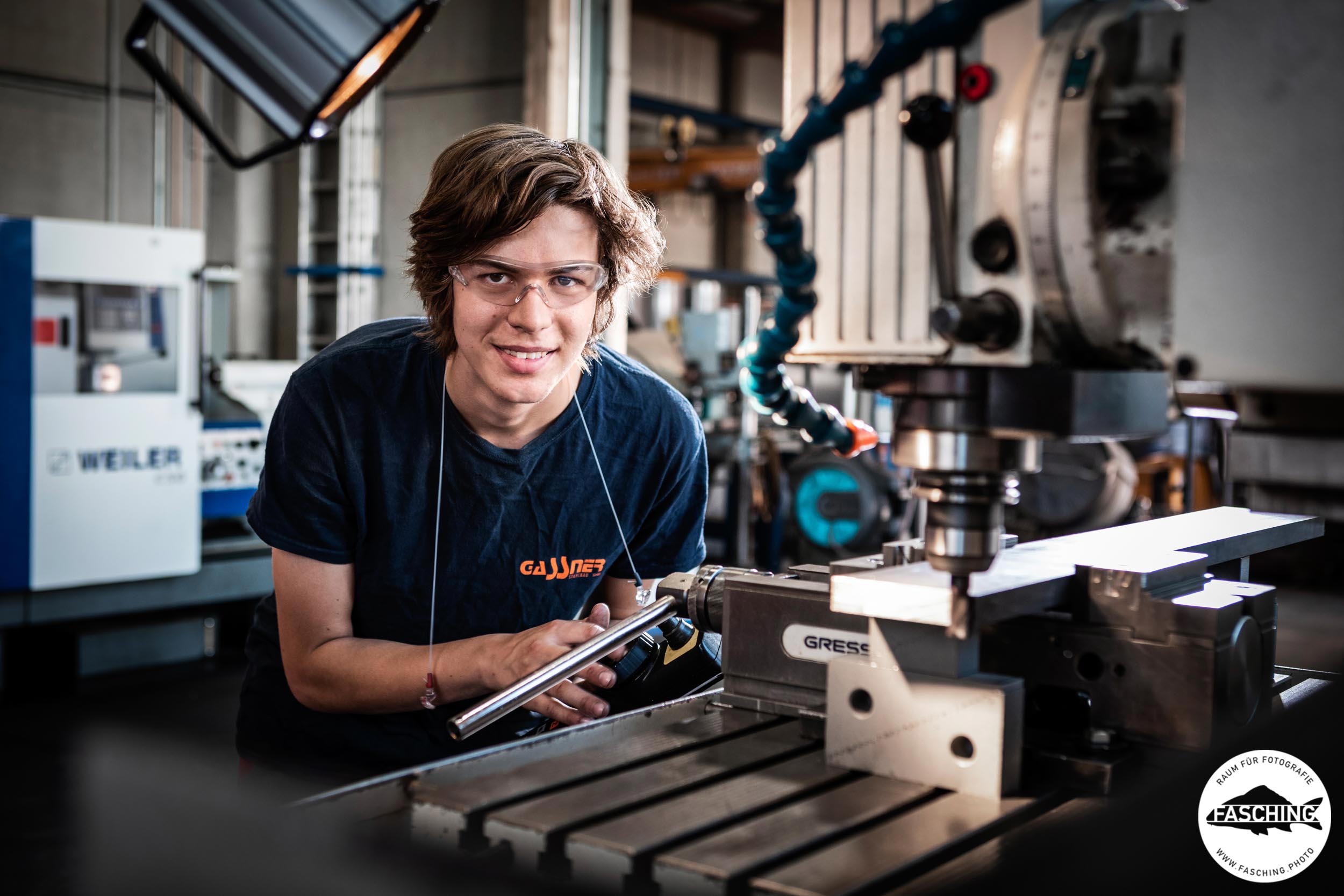Lehrling in dunkelblauem T-Shirt mit Schutzbrille an einer Industriemaschine